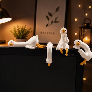 Könnte beinhalten: Vier weiße Gänsefiguren mit orangefarbenen Schnäbeln und Füßen in verschiedenen Posen. Eine liegt flach, eine hängt kopfüber, eine sitzt aufrecht und eine lehnt sich über den Rand. Diese kleinen Dekofiguren sitzen oben auf einem Monitor.