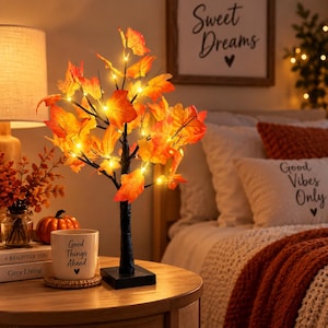 Puede incluir: Una lámpara decorativa de mesa con forma de árbol, con tronco y ramas negras adornadas con hojas de arce de colores naranja, amarillo y rojo. El árbol está iluminado por pequeñas luces LED de color blanco cálido entre el follaje, sobre una base cuadrada.