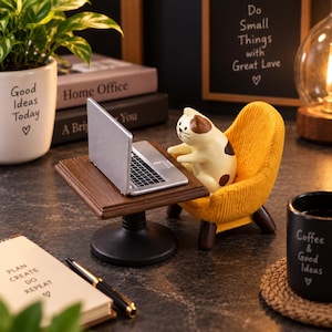 Könnte beinhalten: Eine Miniaturfigur einer cremefarbenen und braunen Katze, die in einem strukturierten senfgelben Sessel an einem kleinen Holztisch mit einem silbernen Laptop sitzt. Der Tisch hat eine dunkelbraune Platte und einen schwarzen Standfuß.