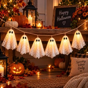 May include: A string of six illuminated ghost-shaped decorations made of white lace fabric. Each ghost features two black oval eyes and glows with a warm light. The garland is suspended horizontally, creating a festive Halloween display.