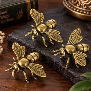 Puede incluir: Tres figuras de abejas de metal en tono latón antiguo con alas detalladas y cuerpos segmentados. Cada abeja está elaborada con un acabado metálico, mostrando patrones de alas intrincados y antenas curvas, presentadas como esculturas decorativas.