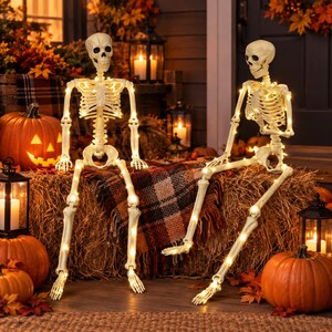 May include: Two off-white plastic skeleton figures in a seated position, each draped with a string of warm white LED fairy lights. The skeletons feature articulated joints and are positioned on a bale of hay, surrounded by orange pumpkins.