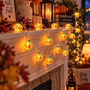 May include: A string of orange pumpkin-shaped LED lights with green stems, illuminated and draped across a white mantelpiece. The lights feature a clear wire and emit a warm glow, creating a festive seasonal display alongside autumn decor.