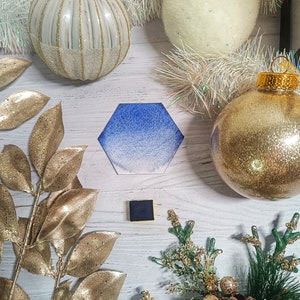 Pode incluir: Uma pintura de aquarela hexagonal azul e branca degradê com uma bandeja de tinta quadrada preta em primeiro plano. A pintura é cercada por ornamentos e decorações de Natal brancas e douradas.