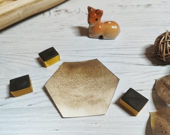 May include: A ceramic deer figurine with brown and white spots sits on a white wooden surface. A hexagonal coaster with a gradient brown finish is surrounded by small, square chocolates with gold wrappers.