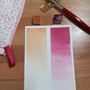 Puede incluir: Dos pastillas de pintura acuarela, una naranja y otra roja, se muestran junto a una hoja de papel blanca con un degradado de pintura acuarela naranja y roja. Un pincel descansa sobre un soporte de pan de oro.
