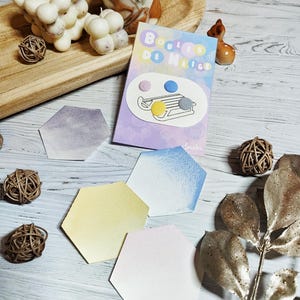 Pode incluir: Uma coleção de itens decorativos, incluindo velas de bolhas, um cartão com o texto "Boules de Nice" e papel aquarela hexagonal em várias cores pastel. Bolas de vime e uma pequena figura de veado também estão presentes.
