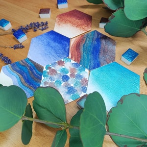 Peut inclure: Un ensemble de six godets de peinture aquarelle hexagonaux avec une variété de couleurs bleues, brunes et sarcelle. Les godets sont disposés en cercle sur une surface en bois avec des feuilles vertes.