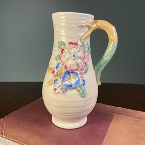May include: Cream-coloured ceramic jug with a textured finish and a branch-like handle. It features floral patterns in pink, blue, and green. The jug is placed on a book.