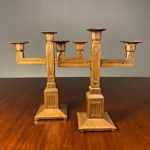 May include: A pair of antique brass candle holders. Each has a square base, a central column, and arms to hold candles. The design features geometric patterns and a warm, aged patina. The candle holders are displayed on a wooden surface.