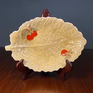 May include: A leaf-shaped ceramic serving dish with a textured surface and scalloped edges. The dish is light yellow with red and green painted accents of tomatoes and leaves. It is displayed on a wooden stand.