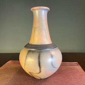 May include: A ceramic vase with a rounded base and a narrow neck. The vase has a light colour scheme with peach and grey tones, and a dark band around the neck. The design includes vertical black streaks.