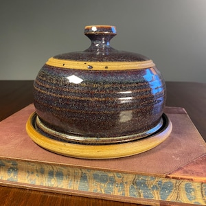 May include: A round ceramic butter dish with a dome-shaped lid featuring a dark purple and blue speckled glaze with horizontal ridges. The base is a contrasting tan colour with a smooth rim. The lid has a small knob handle at the top centre.