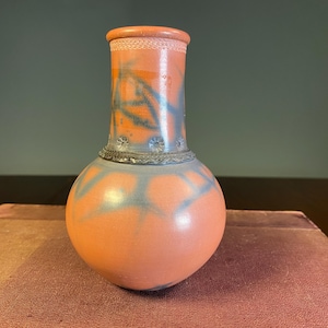 May include: A ceramic vase with a rounded base and a cylindrical neck. The vase is primarily orange with blue-grey abstract designs. The neck has a decorative band and a lighter orange rim. The vase is likely handmade and suitable for home decor.