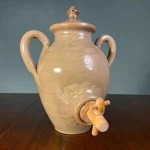 May include: A beige ceramic water jug with a lid, two handles, and a wooden spigot. The jug has a rounded body and a cork around the spigot. The lid has a small knob. The jug is on a wooden surface.