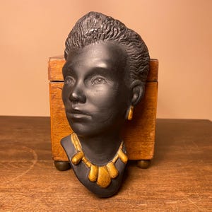 May include: Black ceramic bust of a person with a gold necklace and earrings. The bust is sitting on a wooden box.