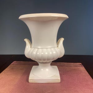 May include: A white urn with a flared top and two curved handles. The urn has a ribbed design around the middle and sits on a square base. The urn is placed on a pinkish-red surface.