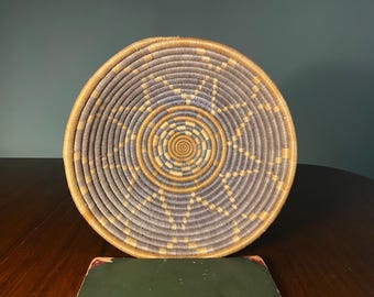 May include: A round woven basket with a spiral design. The basket features a central spiral in shades of tan and blue, surrounded by concentric circles of the same colors. The outer rim is a solid tan color. The basket is on a dark wooden surface.