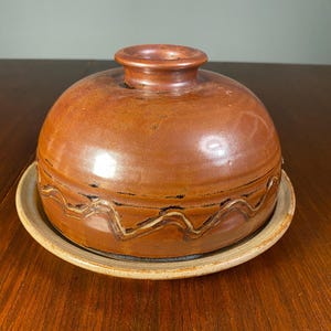 May include: A brown ceramic butter dish with a domed lid and a round base. The lid has a wavy design and a small handle. The dish is glazed with a glossy finish. The dish is likely handmade.