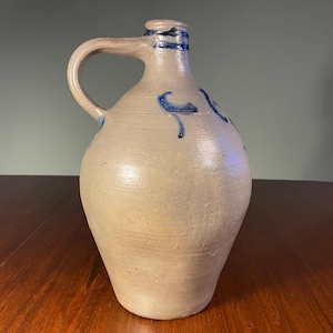 May include: A stoneware jug with a handle and a spout. The jug is a light beige colour with blue painted accents. The jug is sitting on a wooden table. The jug is approximately 30 cm tall.