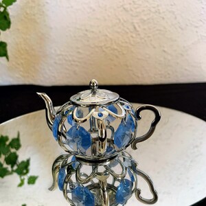 May include: A silver teapot with a decorative design and blue crystal accents. The teapot is sitting on a silver stand.