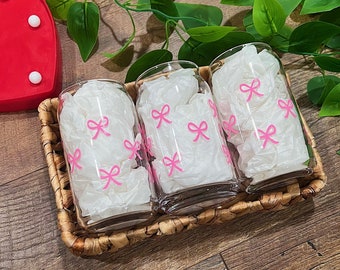 Könnte beinhalten: Drei klare Glasbecher mit pinkfarbenen Schleifen darauf gedruckt. Die Becher stehen in einem geflochtenen Korb.