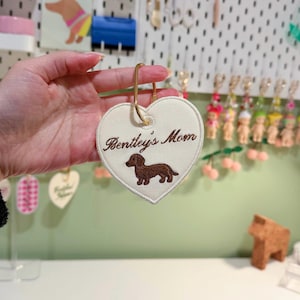 Op de afbeelding: Crème hartvormige sleutelhanger met de geborduurde tekst "Bentley's Mom" en een bruine teckel. De sleutelhanger heeft een goudkleurige metalen ring voor bevestiging.