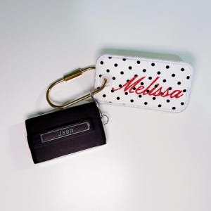 May include: A black Jeep key fob and a white key tag with black polka dots. The key tag has the name "Melissa" embroidered in red thread. A gold-colored carabiner clip connects the two items.