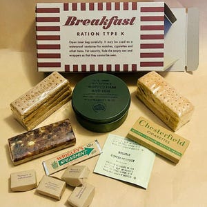 Puede incluir: Un kit de raciones militares vintage etiquetado como "Breakfast, Ration Type K". El kit incluye galletas saladas, una lata de "Chopped Ham and Egg", chicle, cigarrillos, café y otros alimentos pequeños. La caja tiene rayas rojas y blancas.