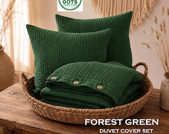 May include: A forest green duvet cover set with a crinkled texture, featuring three visible wooden buttons on the folded edge. The set includes two matching square pillow shams, all displayed in a woven basket with a GOTS certified textile logo.