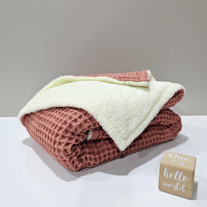 May include: A folded blanket featuring a dusty rose waffle-weave fabric, with a cream-coloured plush lining. The blanket is neatly folded, highlighting the contrasting textures and colours. A wooden block with the text "One day hello world" is in the foreground.
