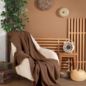 May include: A solid dark brown waffle-textured throw blanket draped over a cream-colored armchair. The blanket features a distinct grid-like pattern across its entire surface, with soft folds cascading down the seat and onto the floor.