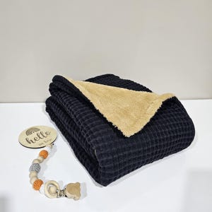 May include: A folded navy blue waffle-weave blanket with a tan sherpa lining. A wooden disc with the text "hello in here" and a rainbow design is next to a wooden and beaded pacifier clip.