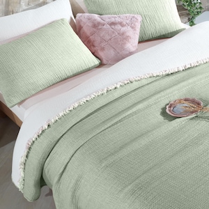 May include: A green waffle-textured throw blanket with a white fringe border. The blanket is folded on a bed with white and green pillows and a pink faux fur pillow.