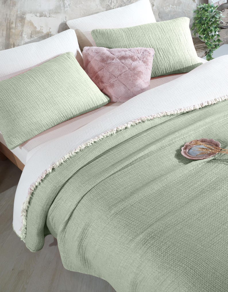 May include: A green waffle-textured throw blanket with a white fringe border. The blanket is folded on a bed with white and green pillows and a pink faux fur pillow.
