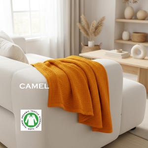 May include: A mustard yellow waffle-textured throw blanket draped over a white sofa. The text CAMEL is printed on the left side, and a circular white and green Global Organic Textile Standard GOTS logo is visible in the lower left corner.