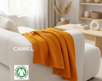 May include: A mustard yellow waffle-textured throw blanket draped over a white sofa. The text CAMEL is printed on the left side, and a circular white and green Global Organic Textile Standard GOTS logo is visible in the lower left corner.