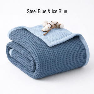 May include: Folded steel blue and ice blue waffle-weave blanket. The blanket is folded in half, with the ice blue side visible on top. A sprig of cotton bolls rests on the blanket. The text "Steel Blue & Ice Blue" is above the blanket.
