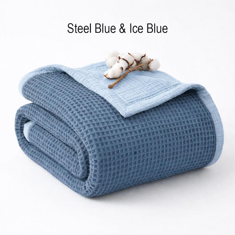May include: Folded steel blue and ice blue waffle-weave blanket. The blanket is folded in half, with the ice blue side visible on top. A sprig of cotton bolls rests on the blanket. The text "Steel Blue & Ice Blue" is above the blanket.