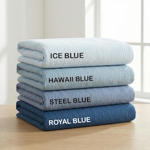May include: A stack of four folded blankets with a textured, crinkled fabric surface. The blankets are labeled with black text as Ice Blue, Hawaii Blue, Steel Blue, and Royal Blue, showing a gradient of blue shades from light to dark.