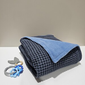 May include: A folded navy blue waffle-weave blanket with a light blue plush underside. A baby rattle with blue star-shaped teethers and a wooden ring is next to the blanket. The blanket is made of soft material.