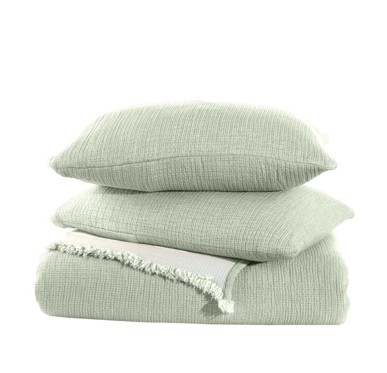 May include: A stack of three sage green waffle weave throw pillows with a matching sage green waffle weave throw blanket. The blanket has a white fringe trim.