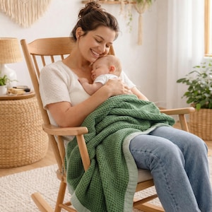 May include: A woman in a white shirt and blue jeans sits in a wooden rocking chair, holding a sleeping baby. A green waffle-weave blanket covers the baby and the woman's lap. The scene is set in a well-lit room.