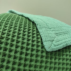 May include: Close-up of a green waffle-weave fabric, possibly a blanket or throw, with a lighter green section folded over it. The texture is visible, showing a grid-like pattern. The colors are muted and natural.