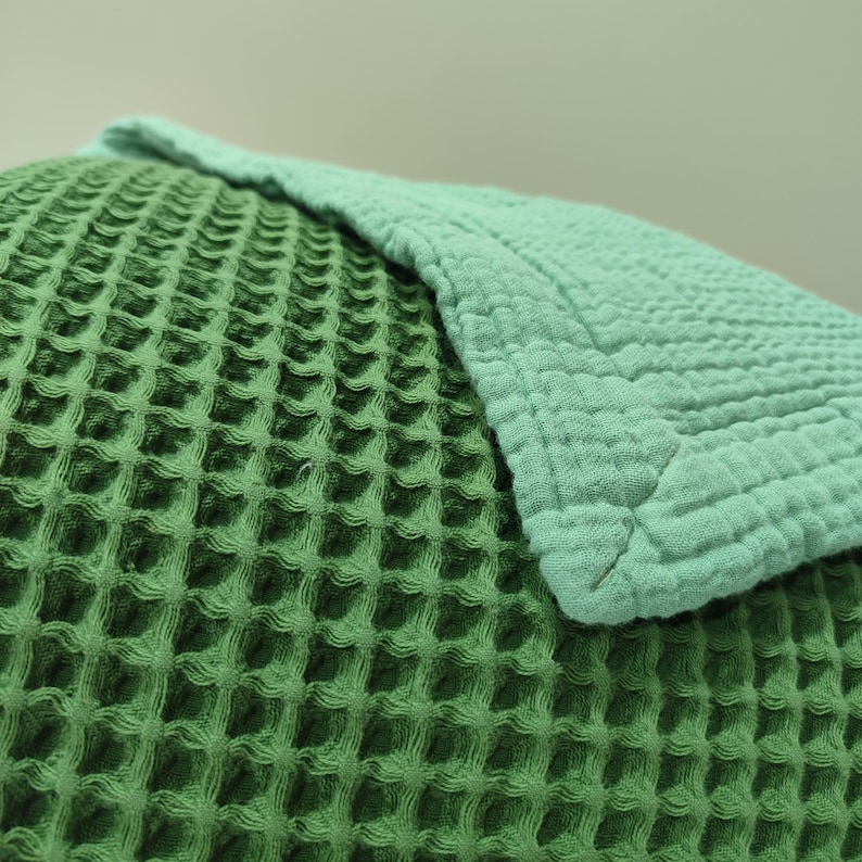 May include: Close-up of a green waffle-weave fabric, possibly a blanket or throw, with a lighter green section folded over it. The texture is visible, showing a grid-like pattern. The colors are muted and natural.