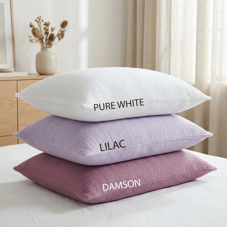 Pode incluir: Tr&ecirc;s almofadas empilhadas em branco puro, lil&aacute;s e ameixa. Cada almofada tem uma superf&iacute;cie texturizada e &eacute; rotulada com o nome da cor em texto branco. As almofadas est&atilde;o em uma cama branca, com um fundo neutro.