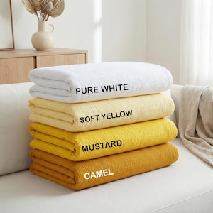 May include: Four folded blankets in shades of white, yellow, and gold are stacked on a white sofa. The top blanket is labeled "PURE WHITE", followed by "SOFT YELLOW", "MUSTARD", and "CAMEL". The blankets are made of a textured fabric.