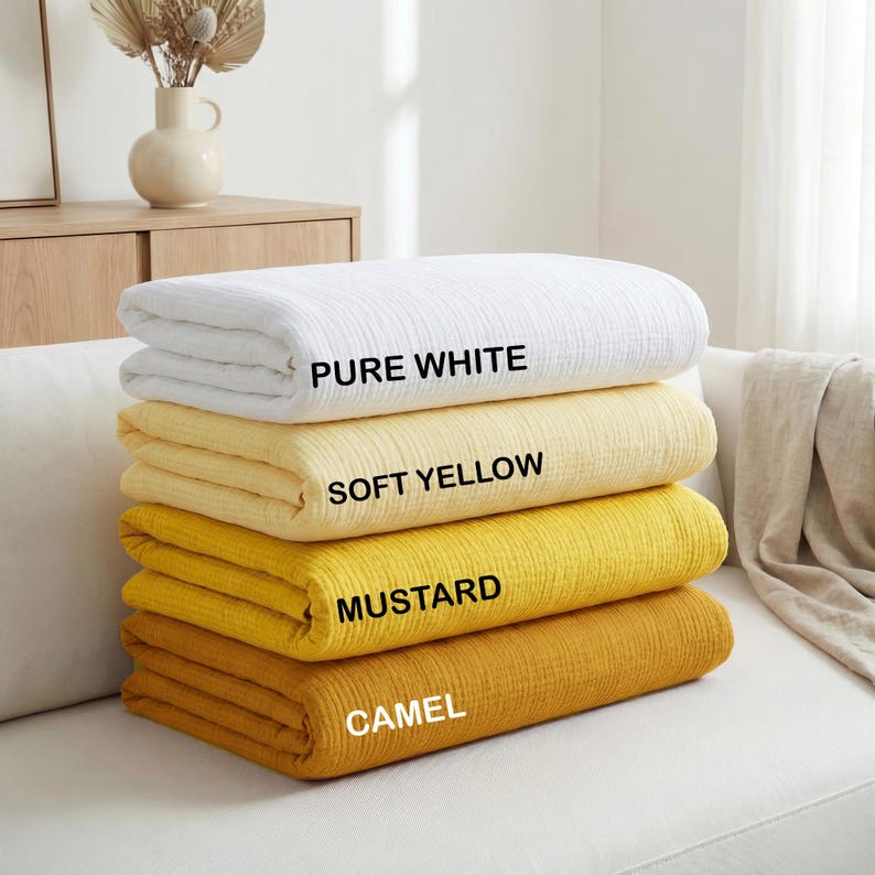 May include: Four folded blankets in shades of white, yellow, and gold are stacked on a white sofa. The top blanket is labeled "PURE WHITE", followed by "SOFT YELLOW", "MUSTARD", and "CAMEL". The blankets are made of a textured fabric.