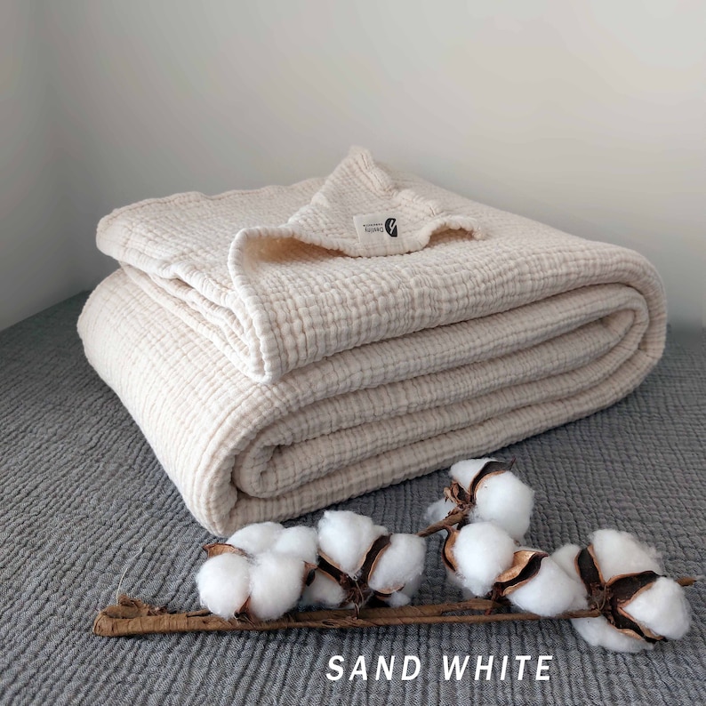 May include: A folded, off-white, waffle-weave cotton throw blanket with a small, white tag that says "Naturally Simple" on a grey textured surface.  The blanket is folded in thirds and has a soft, crinkled texture.  A sprig of cotton bolls is in the foreground.