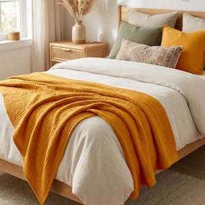 K&ouml;nnte beinhalten: Eine senfgelbe, texturierte Baumwoll-Musselin-Tagesdecke, die &uuml;ber einem Bett mit wei&szlig;em Bettbezug liegt, mit einem Holzbettrahmen, einem Nachttisch aus Rattan und dekorativen Kissen in Olivgr&uuml;n, Senfgelb und einem gemusterten neutralen Farbton.